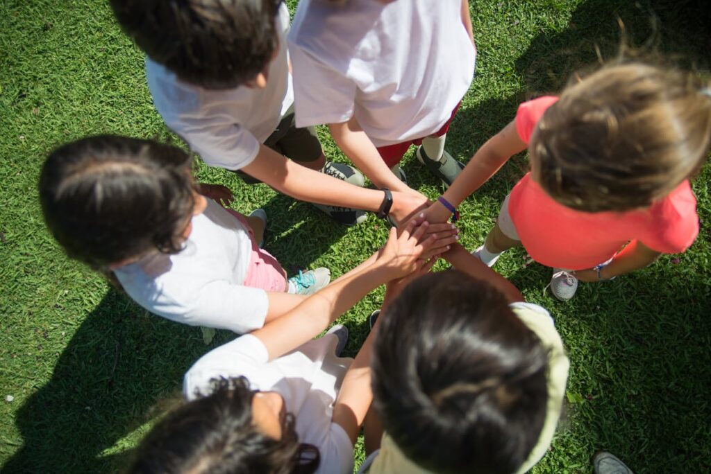 A group of six children stand in a circle on green grass, placing their hands together in the center for a team gesture, viewed from above.